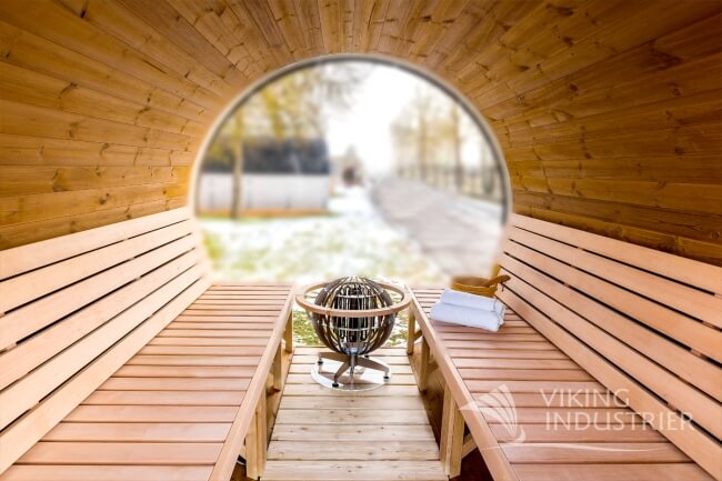 Interior view of a Viking Industrier Barrel Sauna 2.2 x 3m featuring wooden benches and a modern heater, showing a blurred outdoor winter landscape through a large circular glass wall.