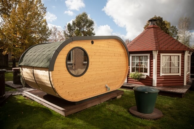 Rear view of the Viking Industrier Delight Camping Pod 2.4 x 4.3m featuring a round window and green shingle roof. Placed on a grassy area with other structures nearby.