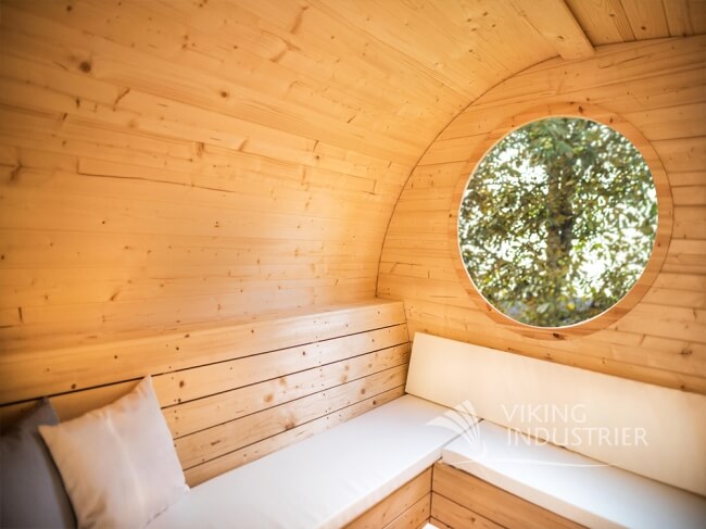 Inside view of Viking Industrier Delight Outdoor Sauna 2.4 x 4.3m with circular window and soft cushions on a wooden background.