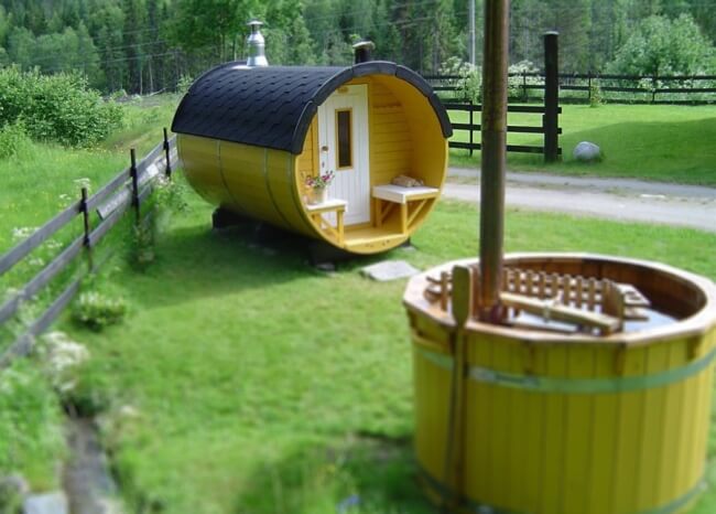 Yellow-painted Viking Industrier Barrel Sauna 2.2 x 3m with black roof and front terrace, located on green grass near a wooden hot tub outdoors.