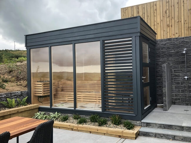 Viking Industrier Sauna Cube with changing room 2 x 4m, exterior view featuring dark gray cladding and full glass panels, placed on a tiled patio with landscaping.