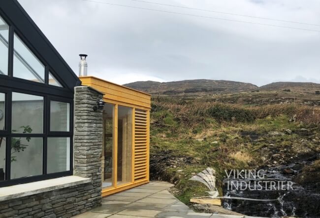 Viking Industrier Sauna Cube with Changing Room 3 x 3m installed beside a house, painted in light wood color with louvered panels, on a natural landscape background.