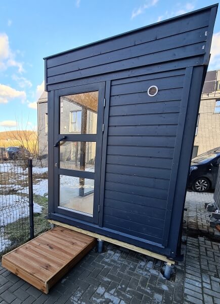 Viking Industrier Sauna Cube 2 x 2m with dark-painted wood exterior and glass door, placed on a small wooden platform beside a paved driveway.