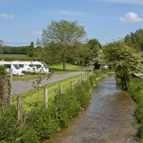 A stream flowing alongside a park with two caravans in, surrounded by trees
