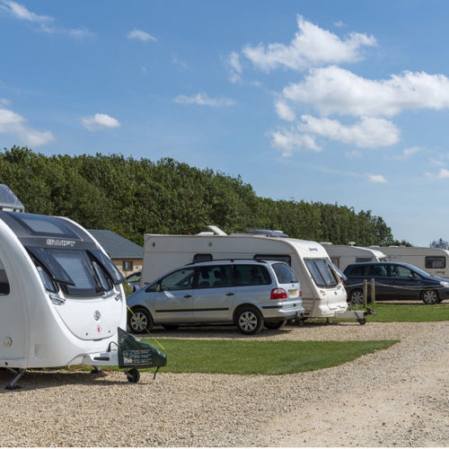 A caravan park with multiple caravans and cars, a dirt track, trees and blue sky in the background