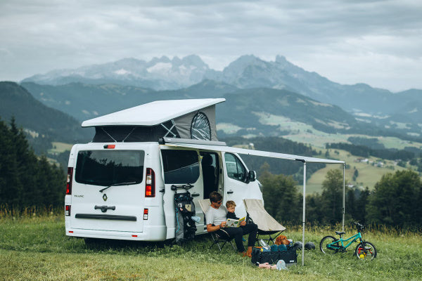 A man sitting under an awning with his child. The awning is connected to a van and the van is in a field with mountains in the background