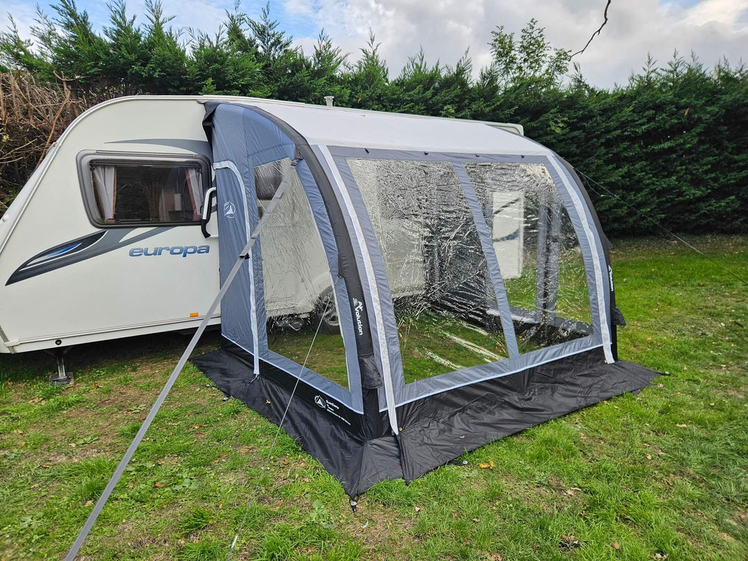 Angled front view of the Sunncamp Ultima Versara Air 260 Deluxe Porch Awning attached to a caravan on grass with hedge trees and cloudy sky.