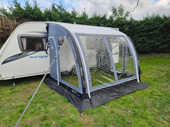 Angled front view of the Sunncamp Ultima Versara Air 260 Deluxe Porch Awning attached to a caravan on grass with hedge trees and cloudy sky.