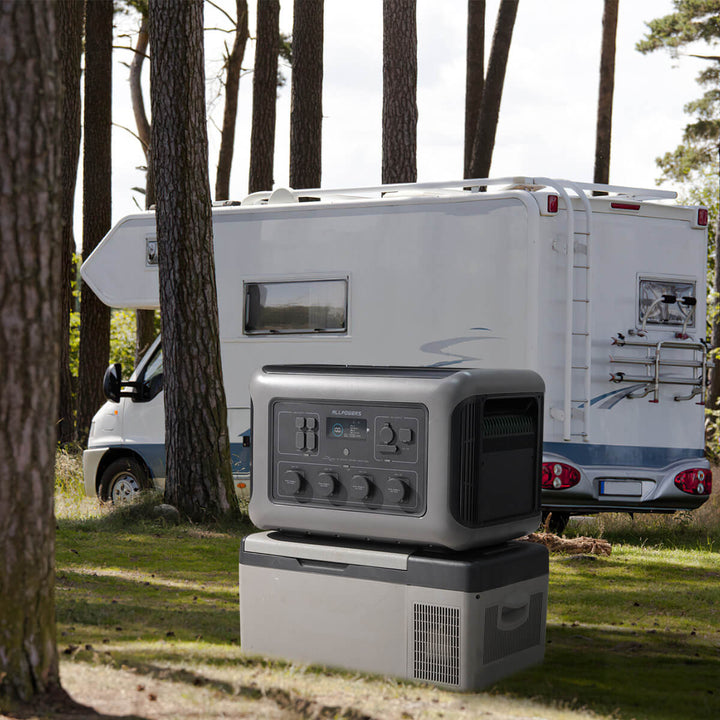 ALLPOWERS R3500 portable power station placed on a portable fridge near a motorhome in a forest campsite.