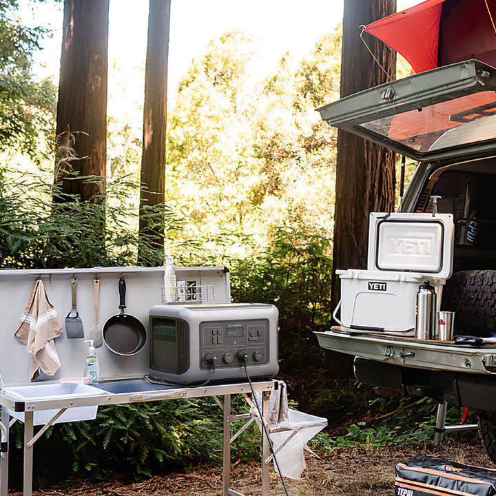ALLPOWERS R3500 portable power station connected at an outdoor campsite beside a vehicle and camping kitchen setup.