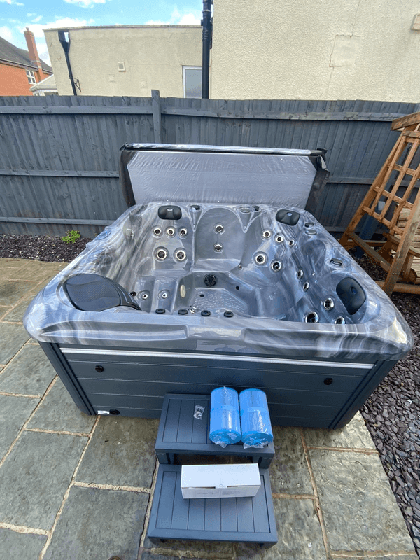 Front view of Luso Spas The Aquarius Plug & Play Hot Tub in grey finish with accessories and filters placed on the steps on an outdoor background.