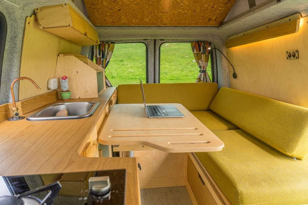 Bivi L Shaped Bed installed in a campervan conversion with overhead cabinets, compact kitchen unit, and countryside view through the windows.