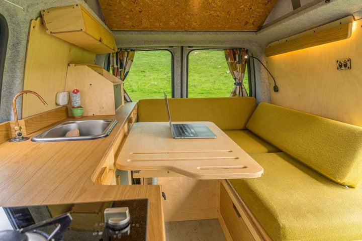Bivi L Shaped Bed installed in a campervan conversion with overhead cabinets, compact kitchen unit, and countryside view through the windows.