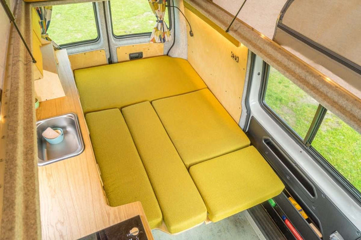 Campervan Bivi L Shaped Bed in daytime setup with L shaped seating, wood kitchen unit, and open rear doors overlooking countryside.