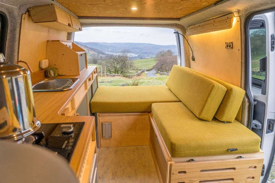 Campervan Bivi L Shaped Bed in mustard yellow upholstery configured as a sofa bed inside a bright campervan interior with wood cabinets and soft lighting.