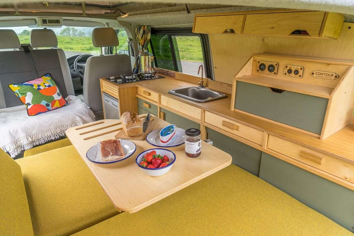Campervan Bivi L Shaped Bed UK version shown as a bed setup with yellow cushions and fitted wood cabinetry inside a campervan.
