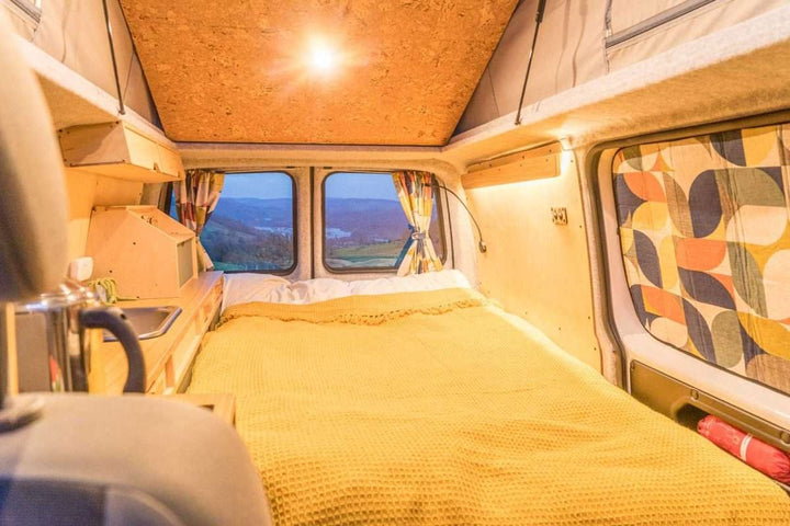 Campervan Bivi L Shaped Bed arranged as a full sleeping platform inside a campervan with wooden kitchen units and scenic countryside visible through the rear doors.