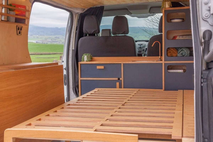 Campervan Navi Kitchen Unit with pull out bed platform extended, blue cabinet fronts and open countryside visible through the doors.
