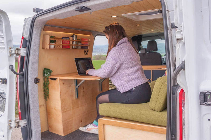 A lifestyle angle of the Campervan Rig Sideboard shows a user seated at the fold-down table, illustrating how the unit functions as a comfortable workstation inside the campervan.