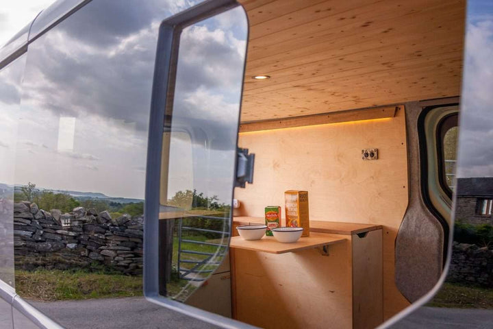 Campervan Rigette Sideboard interior reflected in the van window, showing fold-out table and cabinetry with stone wall and countryside landscape in the background.