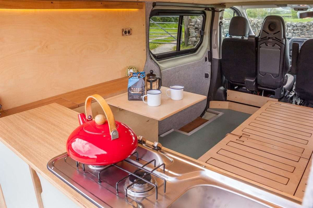 Campervan Rigette Sideboard interior view showing fold-out table, seating area and wooden sideboard unit inside the van, parked outdoors with greenery visible through the windows.