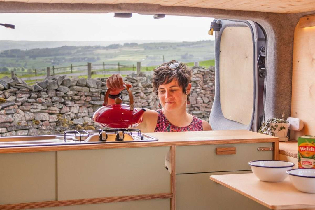 Campervan Rigette Sideboard installed inside a campervan with a woman holding a red kettle over the hob at the open rear doors, with countryside stone wall and rolling hills in the background.