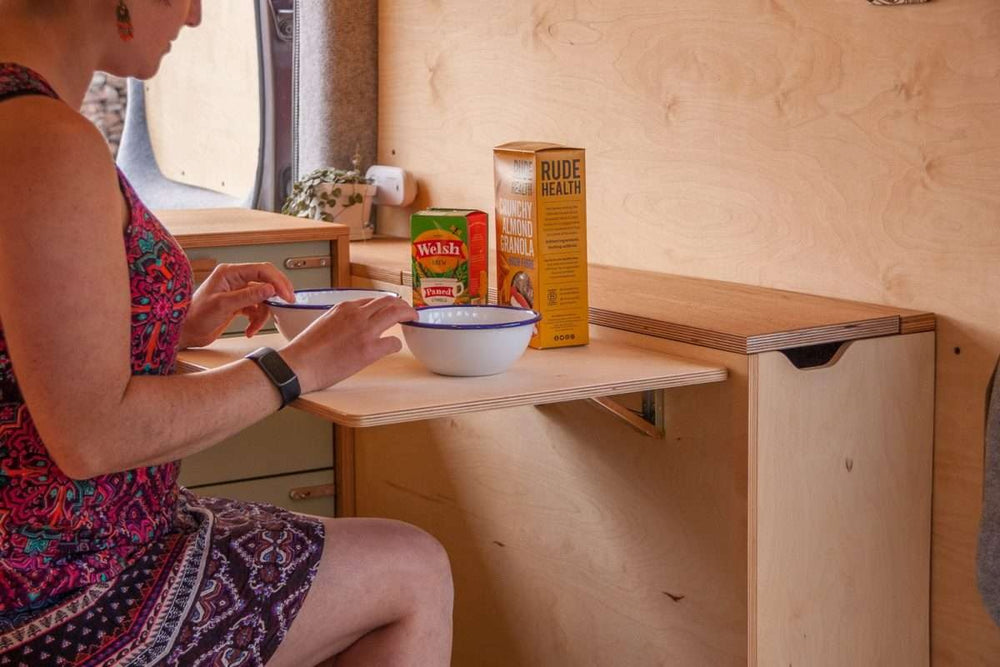 Campervan Rigette Sideboard in use with a woman seated at the fold-out table inside the campervan, with wooden cabinetry and countryside visible through the open doors.