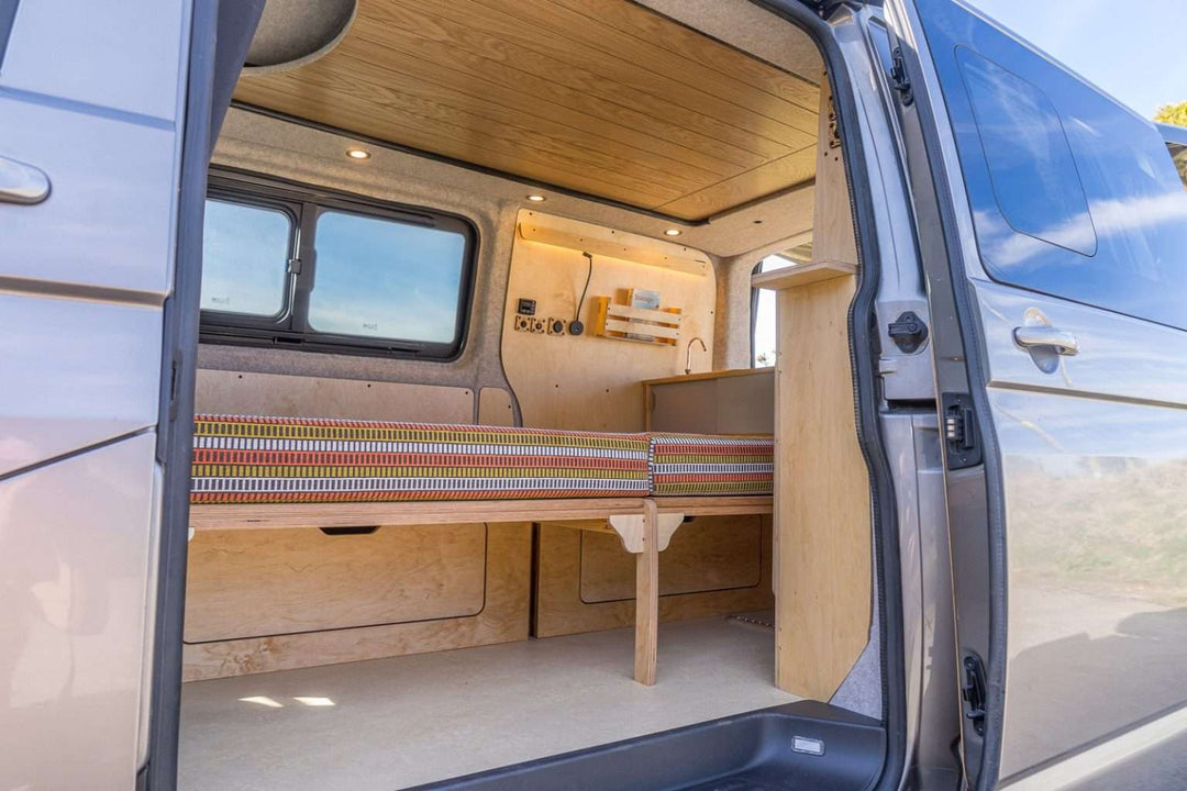 Campervan “Topo” Sofa-Bed in sofa mode with orange cushions beside a wooden kitchen unit, countryside visible through open rear doors in the background.