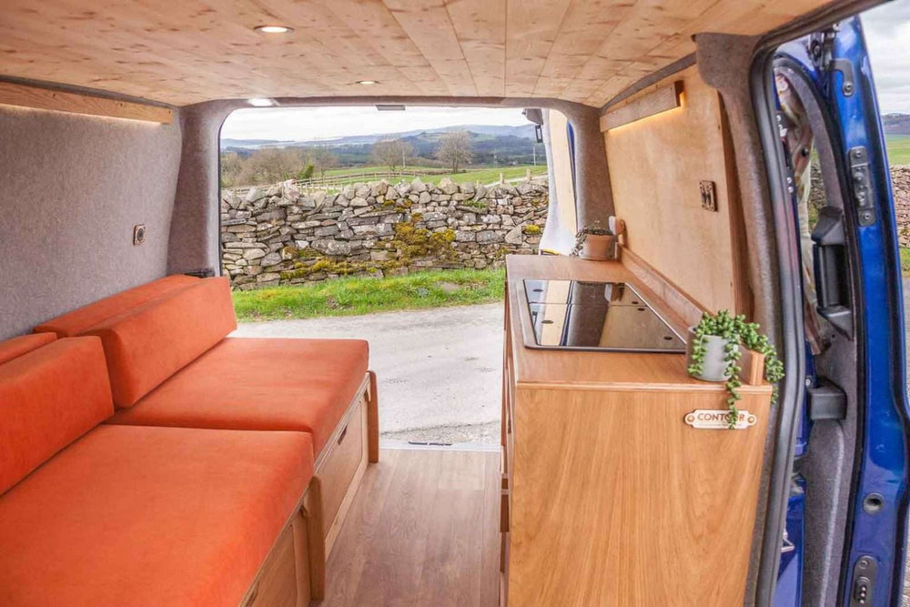Campervan “Topo” Sofa-Bed installed in a campervan with orange bench cushions and wooden kitchen unit, parked outdoors with rural scenery in the background.