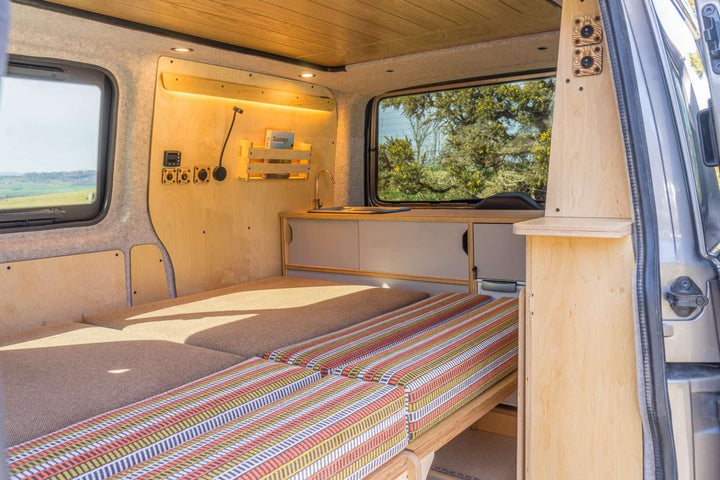 Campervan “Topo” Sofa-Bed set up as a rear sleeping platform with kitchen unit and countryside visible through open doors in the background.