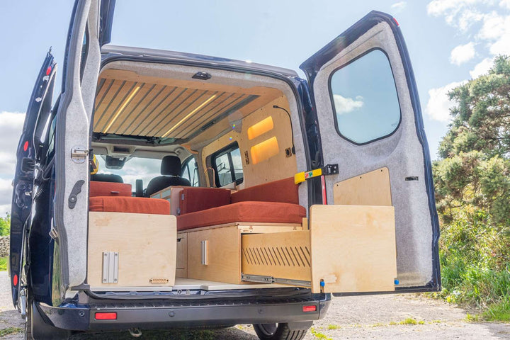 Campervan “Zawn” U-Berth Bench and Bed Unit full rear view with U-shaped seating, pull-out kitchen pod and fold-out table, parked outdoors with rear doors open.