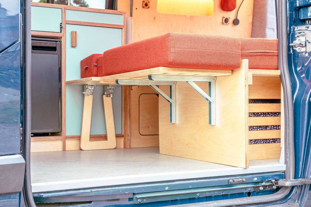 Campervan “Zawn” U-Berth Bench and Bed Unit with pull-out bed platform and storage compartments inside a blue van, shown from rear doors open outdoors.