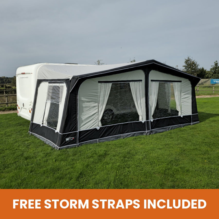 A black and white Camptech Cayman caravan awning is pitched on a green grassy field next to a white caravan. The tent features large window panels with white curtains and a solid black base.