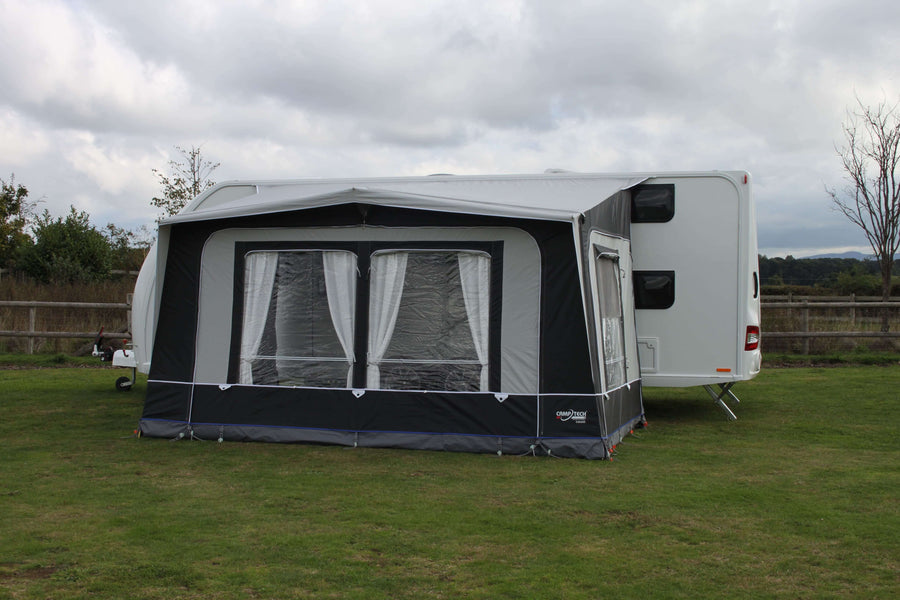 Camptech Count DL porch awning with clear windows and white curtains, attached to a white caravan on green grass under a cloudy sky.