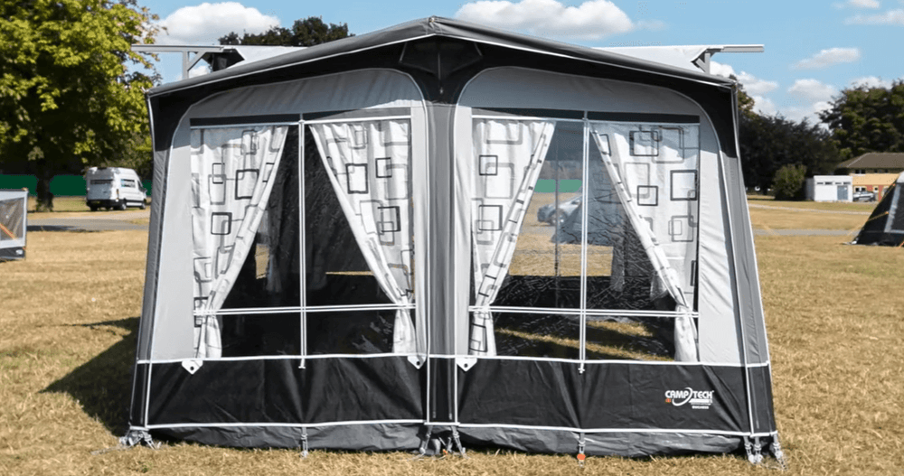 Camptech Duchess porch awning front view showing large clear panels and patterned curtains, set up on dry grass under sunny weather.