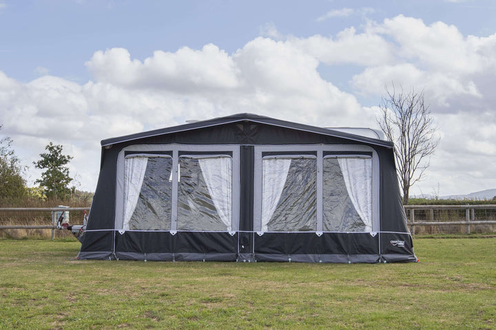 Camptech Hampton DL air awning front view showing clear windows and curtains, pitched on grass beside a caravan under a partly cloudy sky.