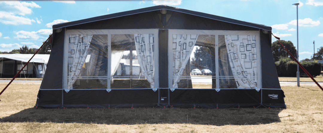 Camptech Kensington Air full caravan awning front view with patterned curtains and large windows, set up on a dry grassy field under a bright blue sky.