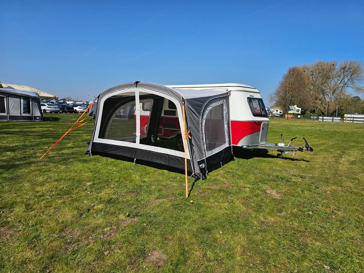 Side view of Camptech Majestic Eriba Caravan Awning set up on a grassy field, connected to a red and white Eriba caravan under a clear blue sky with trees and other caravans in the distance.