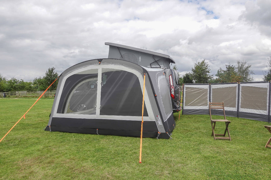 Camptech Moto Gala Driveaway Awning attached to a camper van, set up on a grassy field with folding wooden chairs and a freestanding windbreak under a cloudy sky.