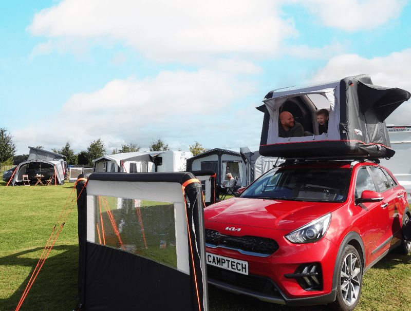 Camptech Queensland inflatable rooftop tent opened on a red car, showing a man and child inside at a campsite with awnings in the background.