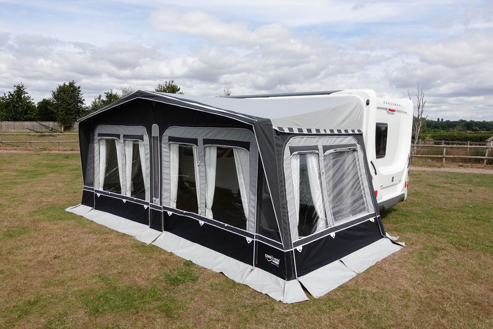 Side view of Camptech Westminster CS seasonal awning with panoramic windows, connected to a white caravan on a grassy campsite with a partly cloudy sky.