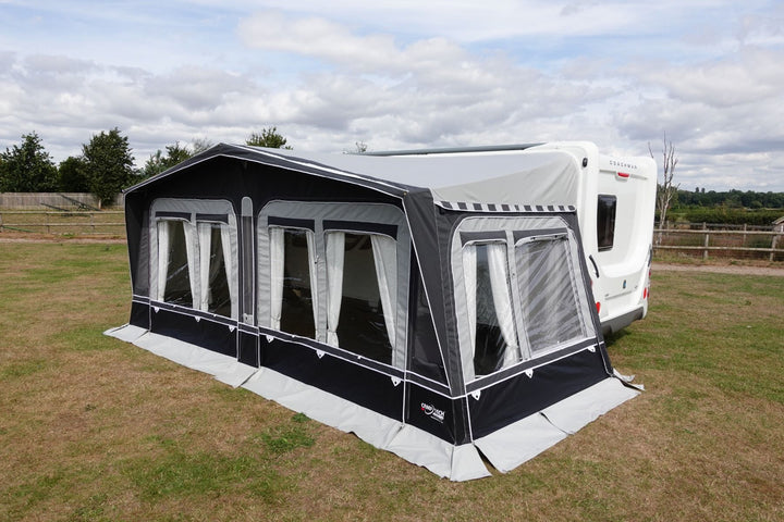 Side view of Camptech Westminster CS seasonal awning with panoramic windows, connected to a white caravan on a grassy campsite with a partly cloudy sky.