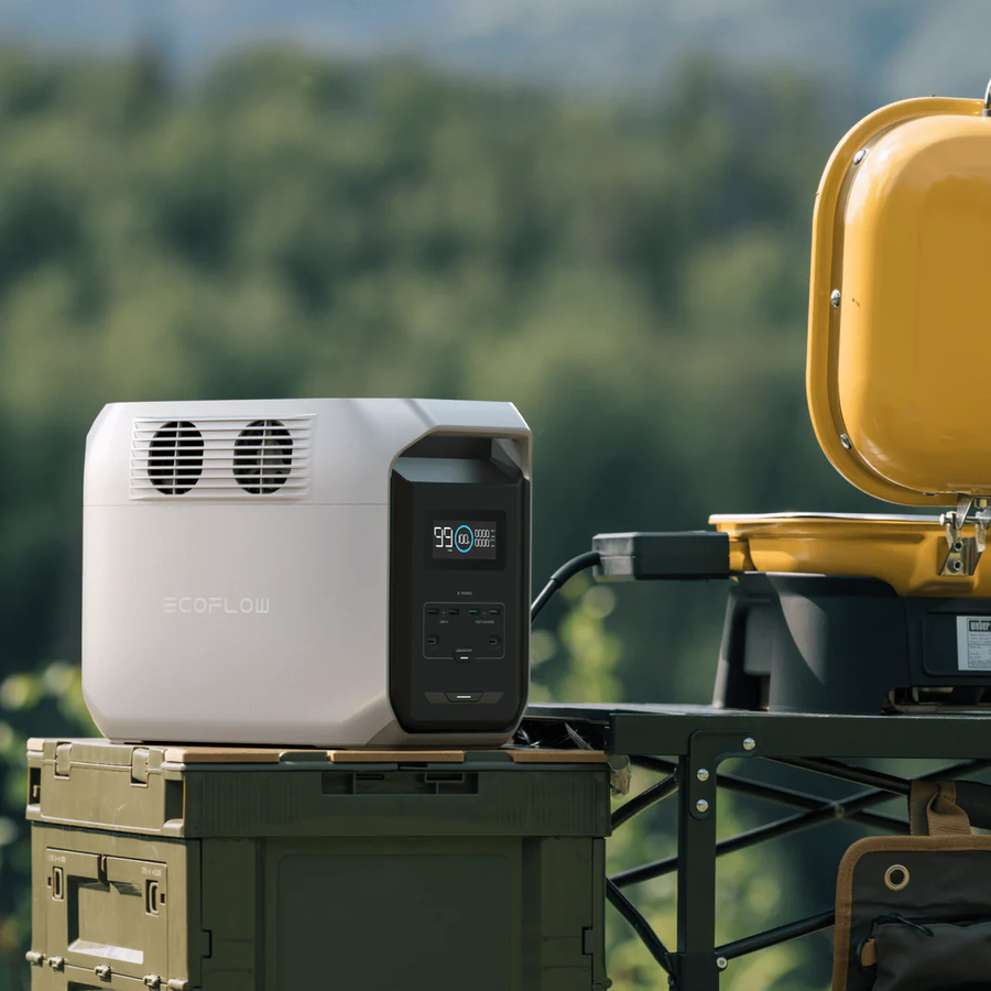 Outdoor scene with the EcoFlow DELTA 3 1500 placed on a green military-style crate, plugged into a yellow electric grill, with a blurred forest background.
