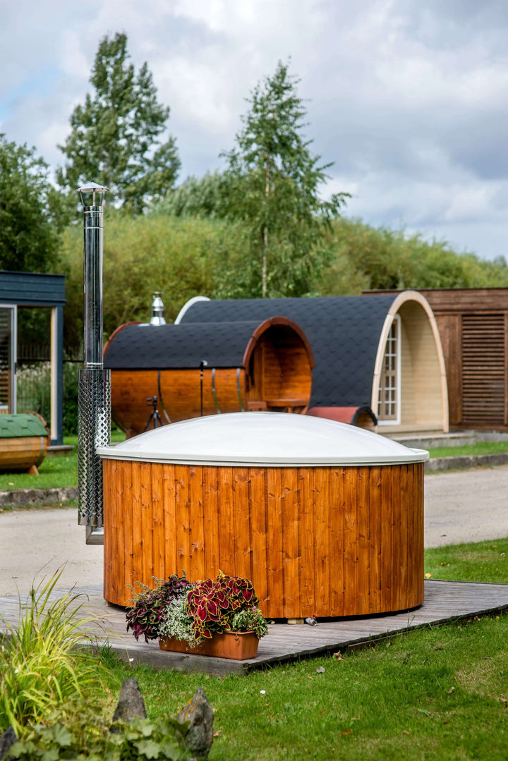 Closed Viking Industrier Fiberglass Hot Tub with integrated heater, standing on a wooden platform with flower pots in the foreground and barrel-shaped wooden saunas in the background under a cloudy sky.