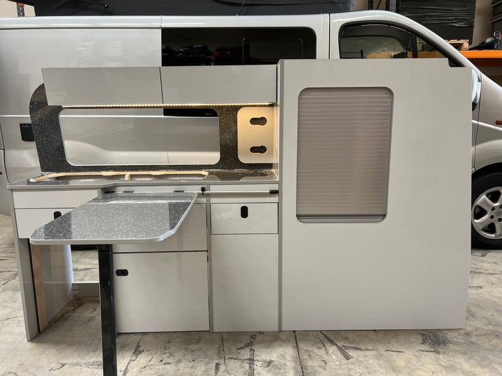 Ford Transit Custom Campervan Kitchen Unit Bespoke installed inside a white van with LED strip lighting, overhead lockers and side tambour door, shown with the van exterior visible in the background.