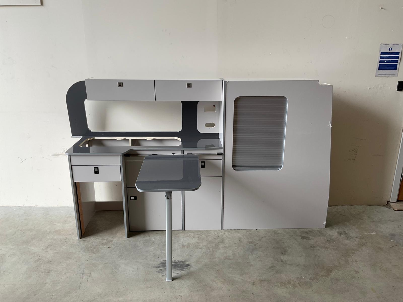 Ford Transit Custom Campervan Kitchen Unit Bespoke in gloss white finish with fold-out table and tambour door, displayed in a workshop on a concrete floor against a cream wall background.