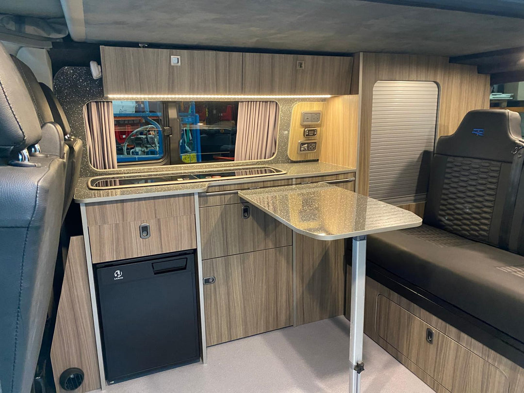 Ford Transit Custom Campervan Kitchen Unit Bespoke in wood-effect finish with integrated fridge and fold-out table, installed inside a campervan with curtains and warm interior lighting.