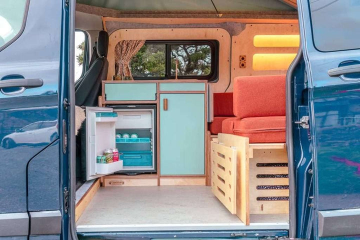 Rear interior view of a campervan showing the Campervan Helm Kitchen Unit with built-in fridge, storage, and seating area.