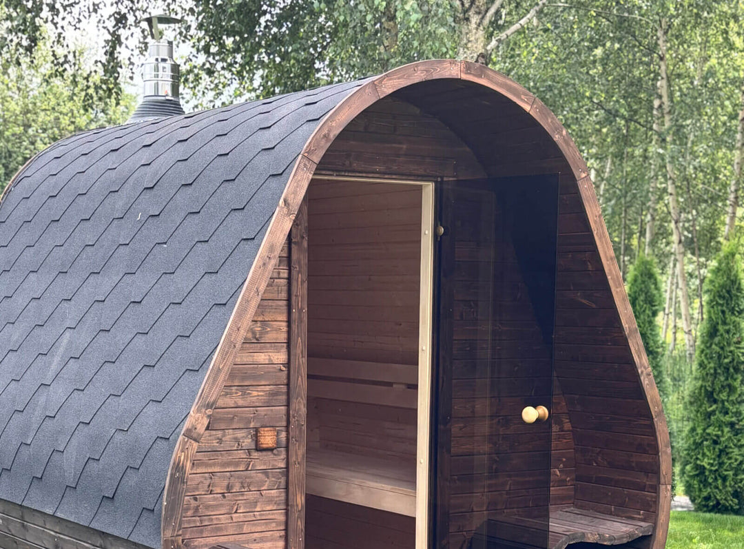 Herkus Grand Triangular Outdoor Sauna exterior close-up with black shingle roof and open glass door, placed in a green garden setting with trees in the background.