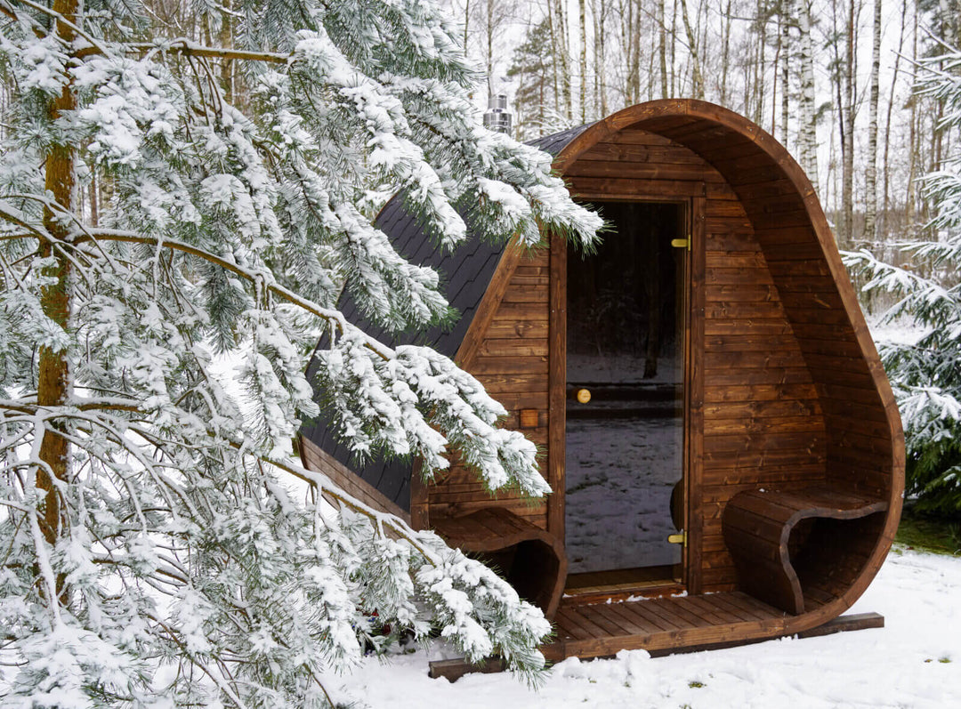 Herkus Grand Triangular Outdoor Sauna front view with dark wood exterior and black shingle roof, surrounded by snow-covered trees in winter.